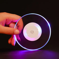 Dessous de verre lumineux tenu de profil dans une main, cercle lumineux bleu-violet allumé sur fond noir.