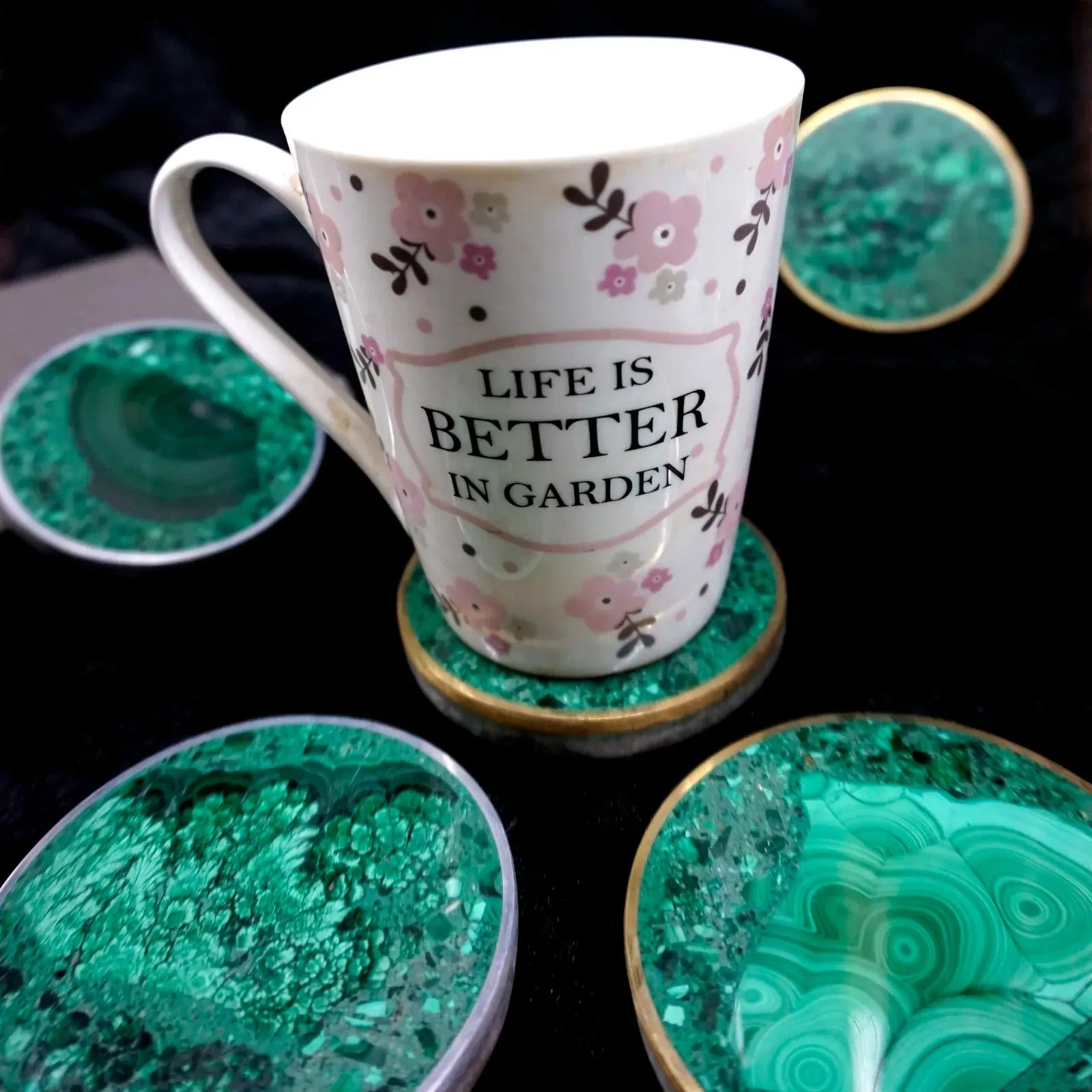 Tasse à motifs floraux posée sur un dessous de verre en malachite verte cerclé, entourée d'autres exemplaires sur fond noir