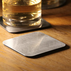 Dessous de verre carré en inox brossé, gros plan sur la texture métallique posée sur une table en bois clair.