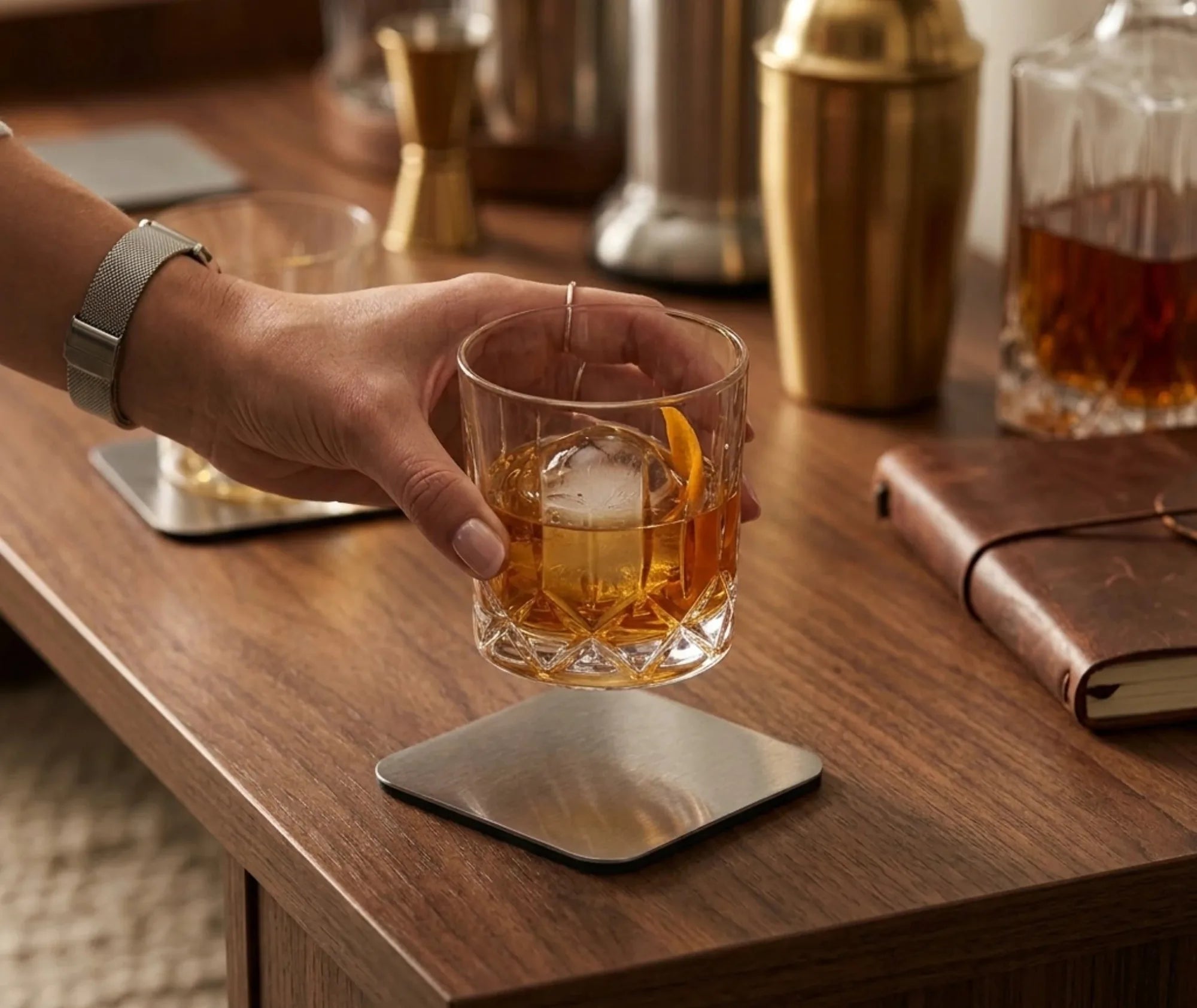 Une main saisit un verre de whisky posé sur un dessous de verre carré en métal, avec shaker et bouteilles de bar en arrière-plan.