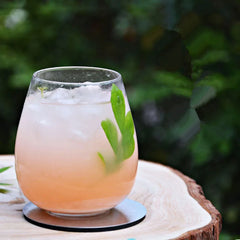 Verre à cocktail rosé garni de menthe posé sur un dessous de verre rond en métal, en extérieur sur une souche en bois.