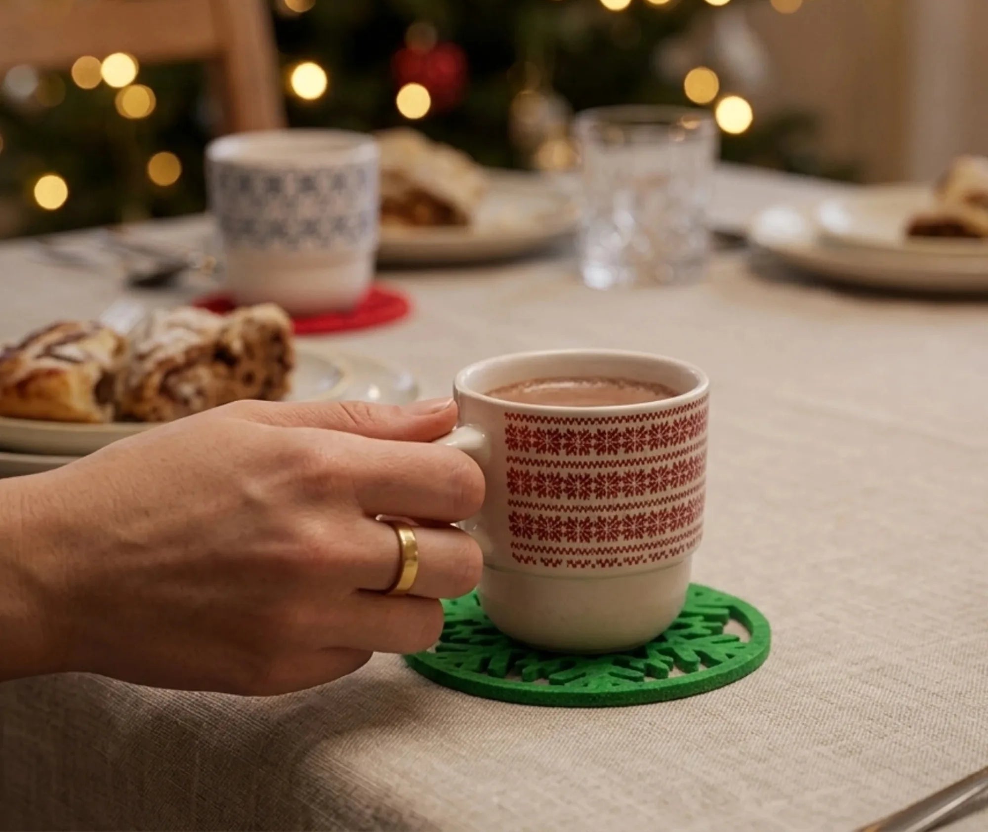 Main tenant une tasse de chocolat chaud posée sur un dessous de verre vert en forme de flocon, sur une table de Noël décorée avec guirlande lumineuse