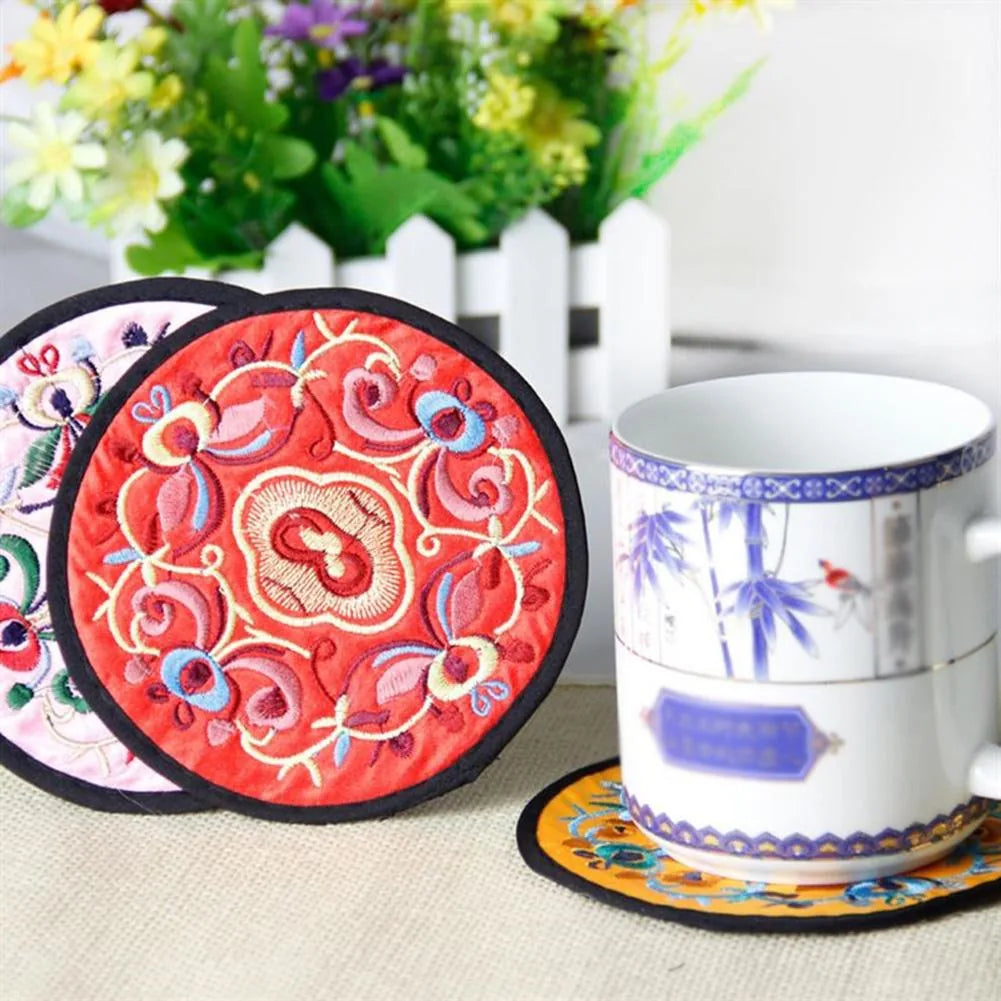 Dessous de verre en tissu brodé rose et rouge appuyés contre une clôture décorative, avec une tasse à motif bambou posée sur un dessous de verre orange.