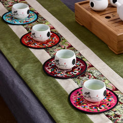 Quatre tasses de thé blanches posées chacune sur un dessous de verre en tissu brodé de couleur différente sur un chemin de table vert.
