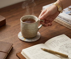 Main tenant une tasse de thé fumante posée sur un dessous de verre vintage, avec un carnet de voyage ouvert à côté.