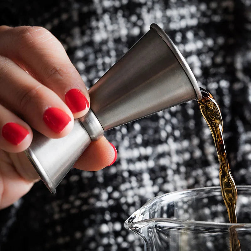 Gros plan sur un doseur à cocktail en inox versant un alcool ambré dans un verre à mélange, tenu par une main aux ongles vernis rouge.