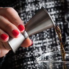 Gros plan sur un doseur à cocktail en inox versant un alcool ambré dans un verre à mélange, tenu par une main aux ongles vernis rouge.