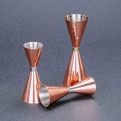 Trois doseurs à cocktail rose gold aux anneaux argenté et doré disposés sur une surface grise texturée