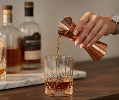 Une main verse du whisky à l'aide du doseur à cocktail cuivré dans un verre à whisky ciselé