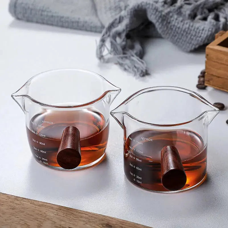 Deux doseurs à cocktail gradués remplis de liqueur ambrée posés sur un comptoir, dans une ambiance cosy avec plaid et grains de café.