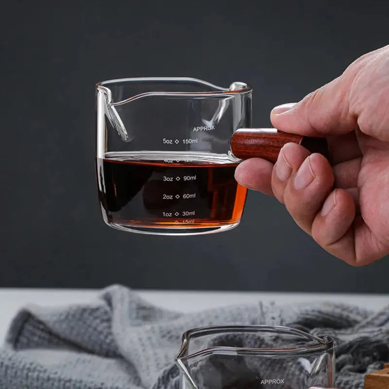 Gros plan sur une main tenant le doseur à cocktail gradué à moitié rempli, laissant apparaître les graduations en onces et millilitres.