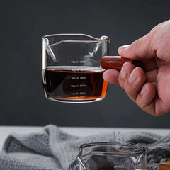 Gros plan sur une main tenant le doseur à cocktail gradué à moitié rempli, laissant apparaître les graduations en onces et millilitres.
