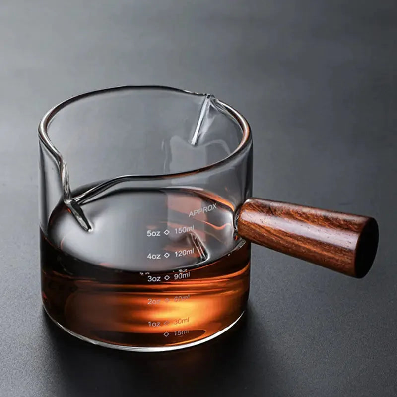 Doseur à cocktail gradué en verre à poignée en bois, à moitié rempli de whisky, sur fond noir dégradé.