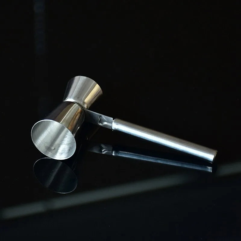 Le doseur à cocktail professionnel en acier inoxydable posé sur une surface noire brillante, avec son reflet visible, dans une ambiance de bar tamisée.