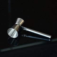 Le doseur à cocktail professionnel en acier inoxydable posé sur une surface noire brillante, avec son reflet visible, dans une ambiance de bar tamisée.