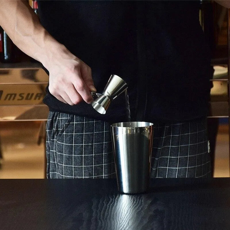 Un barman verse un dosage de liquide depuis le doseur cocktail professionnel dans un shaker en acier inoxydable posé sur un comptoir de bar.