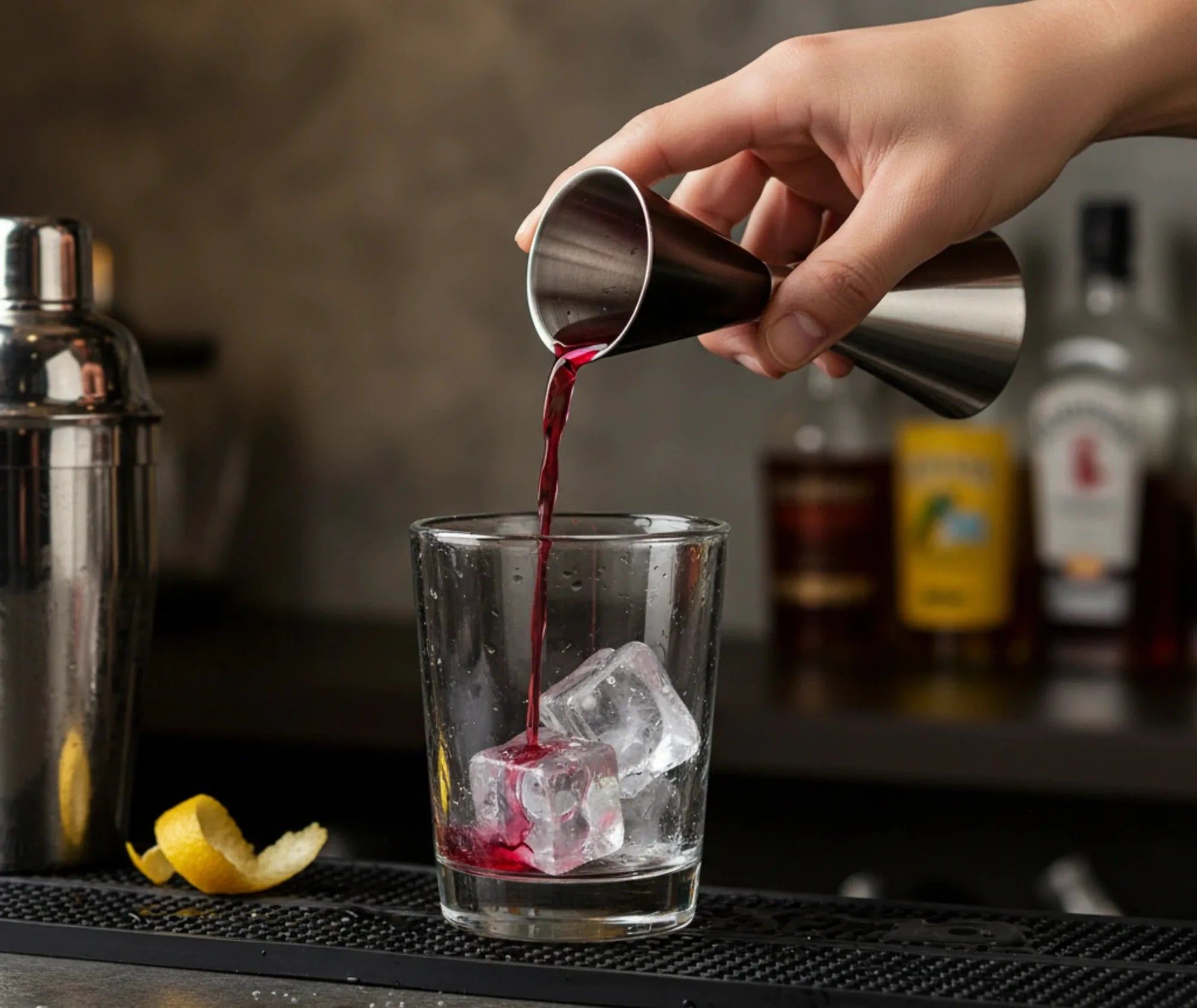 Doseur professionnel versant un cocktail rouge dans un verre avec glaçons et zeste de citron