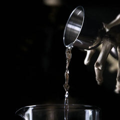 Gros plan artistique d'un doseur à cocktail versant un liquide transparent dans un verre à mélange sur fond noir.