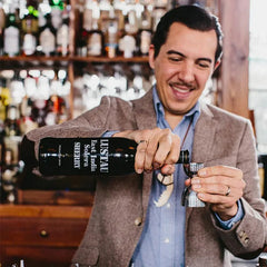 Homme en veste chinée versant une bouteille de sherry dans un doseur à cocktail argenté au comptoir d'un bar.