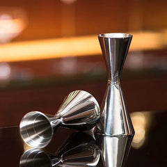 Deux doseurs à cocktail en inox posés sur un comptoir avec reflets, fond de bar flou