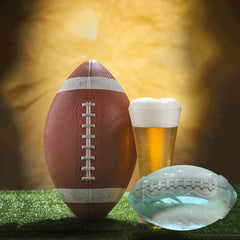 Glaçon transparent en forme de ballon de rugby posé à côté d'un vrai ballon et d'une bière sur un terrain