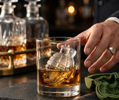 Un glaçon en forme de grenade dans un verre de whisky tenu par une main portant une bague, ambiance chaleureuse de bar avec des carafes en arrière-plan