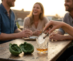 Des amis profitent d'un apéritif en terrasse pendant qu'une pince dépose un glaçon en forme de grenade dans un verre de whisky, à côté du moule à glaçons Grenade