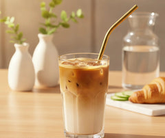 Paille courbée dorée plongée dans un café glacé au lait, posée sur une table en bois avec un livre ouvert.