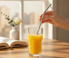 Main tenant une paille courbée en inox plongée dans un verre de jus d'orange, près d'une fenêtre lumineuse.