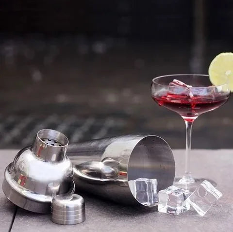 Shaker en inox du kit cocktail en bois ouvert à côté d'un cocktail Cosmopolitan garni de citron vert et de glaçons.