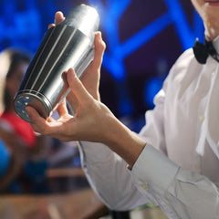 Barman en chemise blanche et nœud papillon tenant un shaker Boston dans une ambiance de bar aux lumières bleues