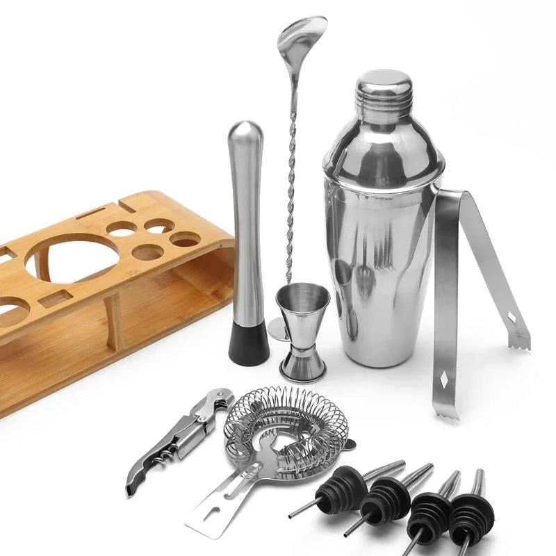 Vue d'ensemble du kit cocktail complet posé sur son présentoir en bambou, avec shaker, pilon, cuillère à mélange, passoire, tire-bouchon, pince à glaçons et becs verseurs.