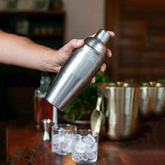 Gros plan d'une main tenant le shaker à cocktail en acier inoxydable au-dessus d'un bar en bois, avec des verres remplis de glaçons à l'arrière-plan.
