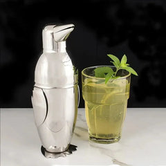 Shaker pingouin en inox posé à côté d'un verre de thé glacé garni de menthe fraîche sur fond noir.