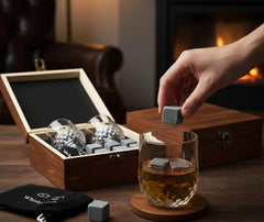 Kit de verres à whisky avec pierres : ambiance cosy près d'une cheminée, une main dépose une pierre à whisky dans un verre rempli de whisky, coffret en bois ouvert avec les verres et les pierres en arrière-plan.
