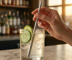 Mini paille en inox plongée dans un gin tonic glacé garni de rondelles de concombre, tenue à la main sur un bar au coucher du soleil.