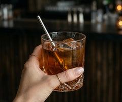 Main ornée de bagues tenant un verre en cristal taillé avec une mini paille en inox dans un cocktail ambré, ambiance tamisée de bar.