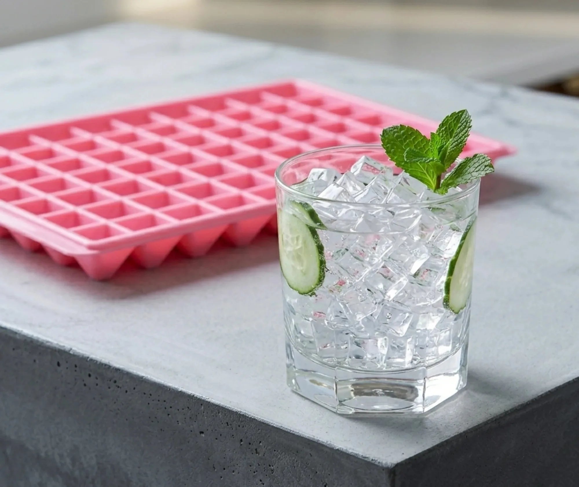 Moule à mini glaçons rose posé à côté d'un verre de cocktail glacé garni de concombre et de menthe