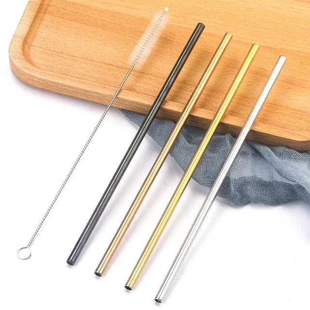 Pailles à cocktail en inox droites de quatre couleurs différentes disposées en diagonale sur une planche en bois clair, à côté d'un linge gris et du goupillon de nettoyage.
