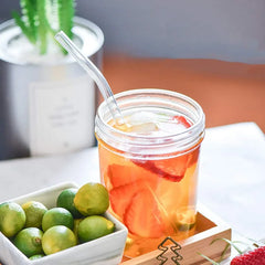 Paille en verre courbée plongée dans un bocal de boisson glacée aux fraises, entourée de citrons verts sur une table.