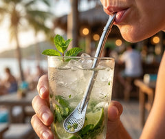 Personne dégustant un mojito à la paille filtrante en inox dans un bar de plage au coucher du soleil