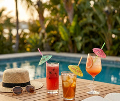 Trois cocktails décorés de pailles à parasol colorées posés sur une table en bois au bord d'une piscine, avec chapeau de paille et lunettes de soleil