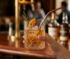 Verre à cocktail old fashioned tenu à la main avec une paille cuivrée, dans une ambiance chaleureuse de bar.