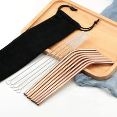 Pailles cocktail cuivrées courbées disposées sur un plateau en bois avec pochette noire et brosses de nettoyage.
