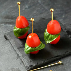 Trois mini-brochettes de tomates cerises et basilic montées sur pics à cocktail dorés, présentées sur une ardoise noire.