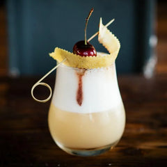 Pique à cocktail doré naturel surmonté d'une cerise, posé sur un cocktail crémeux et mousseux.