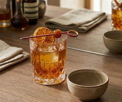Pique à cocktail rouge orné d'une rondelle d'orange séchée et d'une cerise, piqué dans un old fashioned ambré.