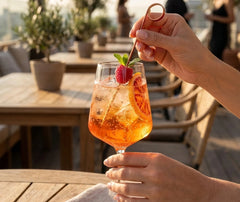 Main plaçant un pique à cocktail orné de framboise et de menthe dans un spritz pétillant en terrasse.