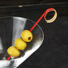 Pique à cocktail rouge coquelicot garni de trois olives vertes, posé sur un verre à martini.