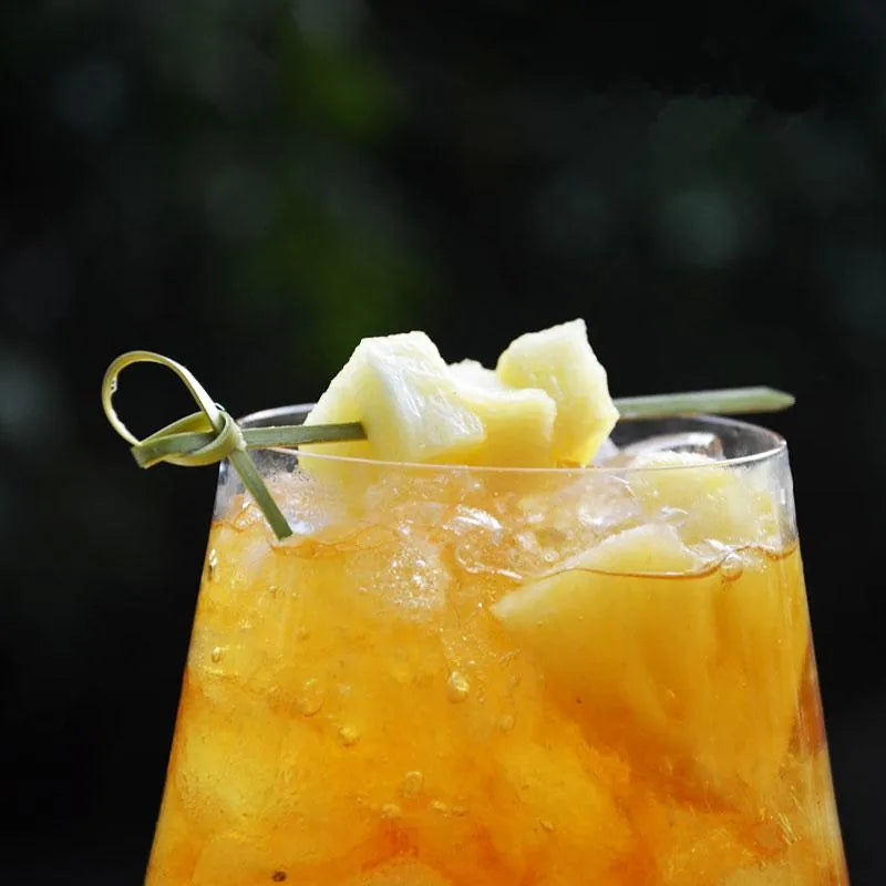 Pique à cocktail vert jungle garni de morceaux d'ananas frais posé sur un cocktail orangé glacé.